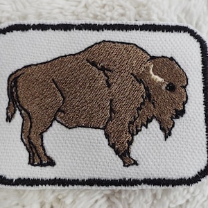 May include: Embroidered patch featuring a brown bison on a white background. The bison is detailed with textured stitching, and the patch has a black border. The patch is rectangular with rounded corners.