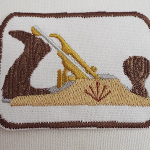 May include: A brown embroidered patch depicting a wood plane with a gold blade and a brown handle. The plane is on a beige background with a red starburst design.