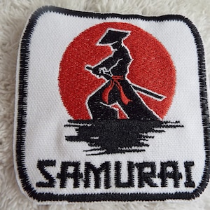 May include: A white embroidered patch with a black silhouette of a samurai warrior holding a sword in front of a red rising sun. The text "SAMURAI" is embroidered in black below the image.