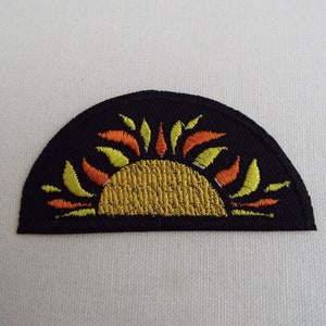 May include: A black fabric patch with an embroidered yellow sun with orange rays. The sun is half-circle shaped and the rays are stylized.