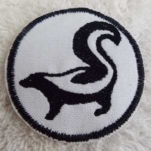 Könnte beinhalten: Ein weißer runder Patch mit einem schwarzen gestickten Rand und einer schwarzen Silhouette eines Stinktiers mit einem weißen Streifen auf dem Rücken.