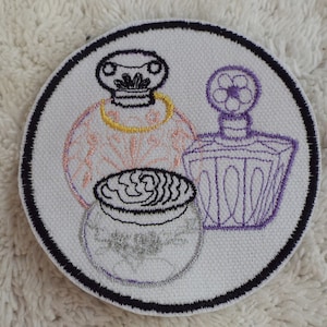 Puede incluir: Parche bordado con tres botellas de perfume. Las botellas están delineadas en negro y rellenas de diferentes colores, incluyendo morado, rosa y amarillo. El parche es circular y tiene un borde negro.