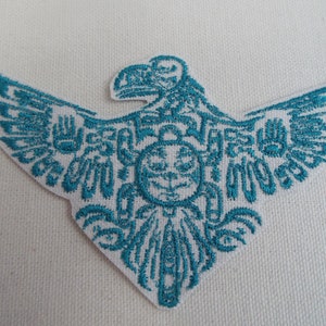 Könnte beinhalten: Türkiser gestickter Aufnäher eines Adlers mit ausgebreiteten Flügeln. Der Adler ist mit geometrischen Mustern und einem zentralen kreisförmigen Design gestaltet.