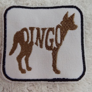 May include: A white embroidered patch with a brown outline of a dog. The dog's silhouette is formed by the word "DINGO" in brown embroidery.
