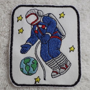 May include: An embroidered patch featuring a cartoon astronaut in a blue spacesuit floating in space with a small planet. The astronaut is holding a rope attached to the planet. There are yellow stars in the background.