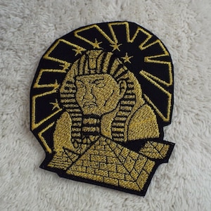 Puede incluir: Parche de hierro bordado en oro con la cabeza de un faraón y una pirámide en el fondo. El parche tiene un fondo negro con un diseño de sol radiante en oro.