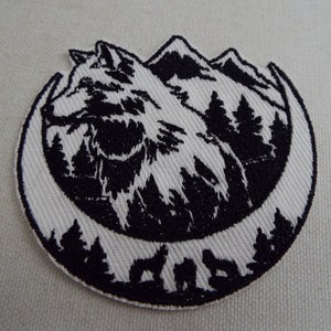 Puede incluir: Parche bordado en blanco y negro con un lobo aullando a la luna con montañas y árboles en el fondo.