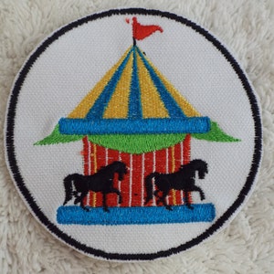 May include: A round, white embroidered patch with a black border. The patch features a colorful carousel with a red, yellow, and blue striped tent, a green base, and two black horses.