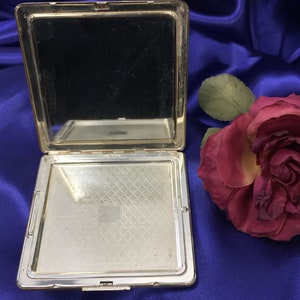 Op de afbeelding: Een zilveren compact spiegel met een scharnierend deksel en een gespiegelde binnenkant. De compact is open en de binnenkant is zichtbaar. De compact ligt op een blauw satijnen oppervlak.