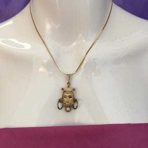 May include: A gold-toned necklace featuring a pendant with a painted face design. The pendant has a cream-colored face with red lips and black eyes, adorned with a crown and earrings. A small pearl is at the bottom of the pendant.