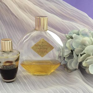May include: Two glass perfume bottles with gold-toned caps. The smaller bottle is tapered with dark liquid and the word WORTH embossed. The larger round bottle contains amber liquid, a diamond-shaped label reading EAU DE TOILETTE JE REVIENS WORTH PARIS.