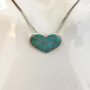 May include: A silver necklace with a heart-shaped pendant. The pendant is filled with turquoise and blue-green mosaic-style stones. The chain is a thin, flat, silver link style.