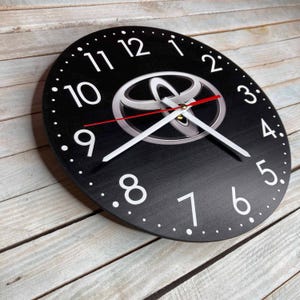 May include: Black round wall clock with white numbers and hands. The clock features a silver Toyota logo in the center.