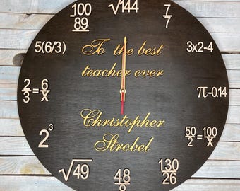 Puede incluir: Un reloj redondo de madera marrón oscuro con números y ecuaciones dorados alrededor del borde. La esfera del reloj tiene el texto "To the best teacher ever" y el nombre "Christopher Strobel" en letras doradas. Las manecillas del reloj son doradas y rojas.
