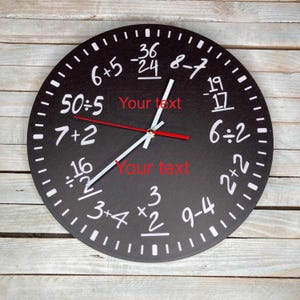 May include: A black, round wall clock with white numbers and equations. The clock face features equations such as 6+5, 50÷5, and 7+2. The clock hands are white with red tips. The background is a light wood.