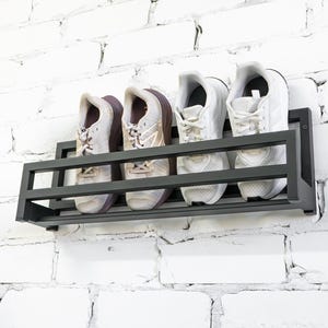 May include: Black metal shoe rack mounted on a white brick wall with four pairs of white sneakers on the rack