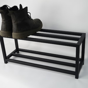 A black metal tier shoe rack. The shoe rack has a simple design with straight lines and a rectangular shape. Two pairs of black boots are on the top shelf.