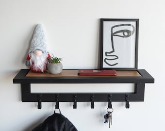 May include: Black metal wall shelf with hooks, a wooden top, and a red book. A small grey gnome and a potted plant are on the shelf. A black and white line art print of a face is hanging on the wall above the shelf.