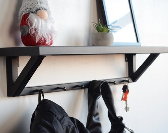 Puede incluir: Un estante de pared de metal negro con una superficie superior plana y una barra horizontal inferior equipada con cinco ganchos espaciados uniformemente. El estante está montado en una pared y sostiene una figura de gnomo, una planta pequeña y un marco negro.