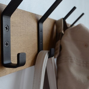 K&ouml;nnte beinhalten: Ein Wand-Kleiderst&auml;nder aus Holz mit drei schwarzen Metallhaken. Eine wei&szlig;e Tasche und ein beiger Hut h&auml;ngen an den Haken.