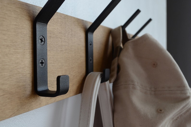 K&ouml;nnte beinhalten: Ein Wand-Kleiderst&auml;nder aus Holz mit drei schwarzen Metallhaken. Eine wei&szlig;e Tasche und ein beiger Hut h&auml;ngen an den Haken.
