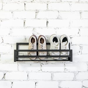 May include: A black metal shoe rack mounted on a white brick wall. Four white sneakers are displayed on the rack.