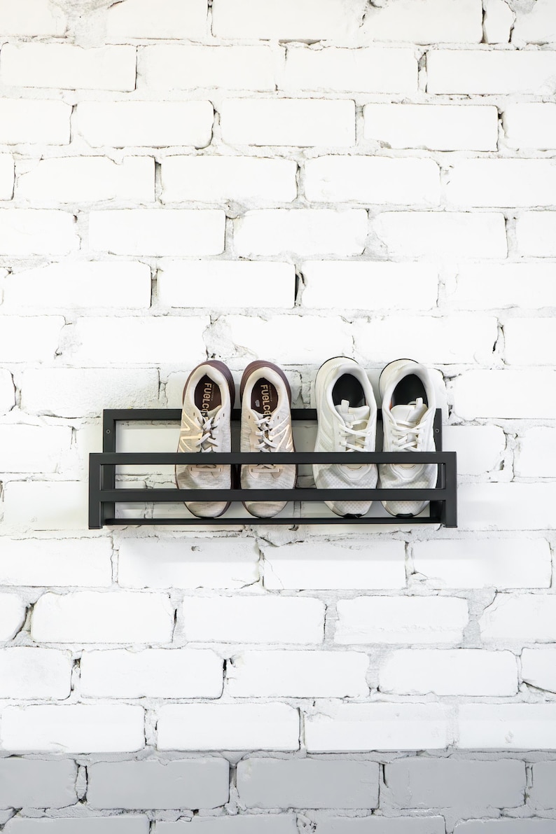 May include: A black metal shoe rack mounted on a white brick wall. Four white sneakers are displayed on the rack.