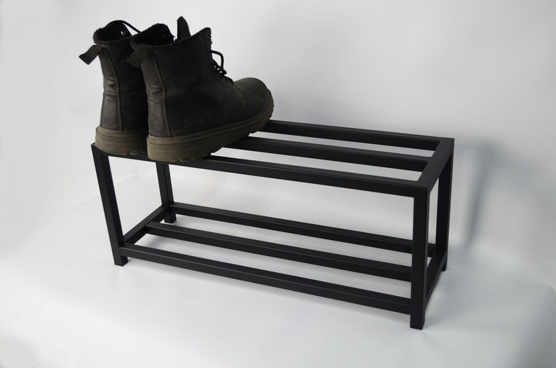 A black metal tier shoe rack. The shoe rack has a simple design with straight lines and a rectangular shape. Two pairs of black boots are on the top shelf.