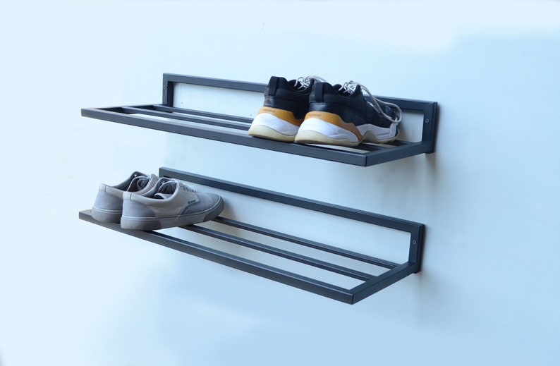 May include: Two black metal shoe shelves mounted on a white wall. The top shelf has two black and white sneakers on it. The bottom shelf has two gray sneakers on it.