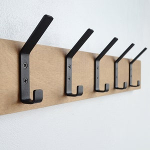 K&ouml;nnte beinhalten: Ein Wand-Kleiderst&auml;nder aus Holz mit f&uuml;nf schwarzen Metallhaken. Der Kleiderst&auml;nder ist aus hellbraunem Holz und die Haken sind mit Schrauben befestigt.