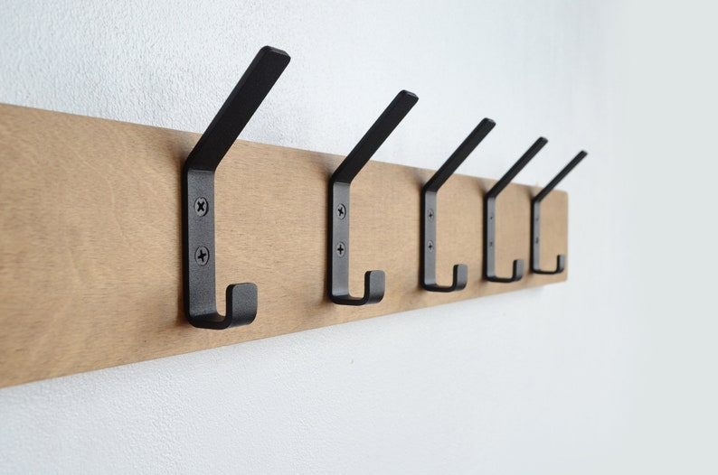 K&ouml;nnte beinhalten: Ein Wand-Kleiderst&auml;nder aus Holz mit f&uuml;nf schwarzen Metallhaken. Der Kleiderst&auml;nder ist aus hellbraunem Holz und die Haken sind mit Schrauben befestigt.