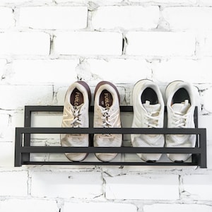 May include: A black metal shoe rack mounted on a white brick wall. Four white sneakers with brown accents are displayed on the rack. The sneakers have the text "FuelCell" printed on the side.