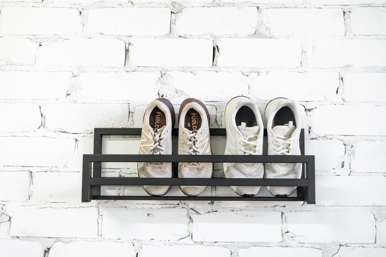 May include: A black metal shoe rack mounted on a white brick wall. Four white sneakers with brown accents are displayed on the rack. The sneakers have the text "FuelCell" printed on the side.