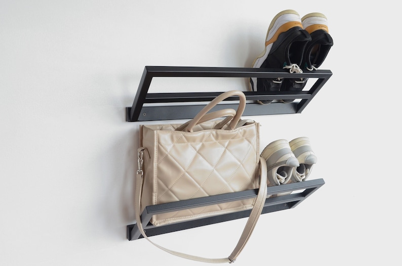 May include: Black metal wall mounted shoe rack with two shelves. A beige quilted handbag with a long strap is hanging on the lower shelf. A pair of white sneakers and a pair of black sneakers with yellow accents are on the top shelf.