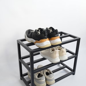 May include: Black metal shoe rack with three shelves, holding six pairs of shoes. The top shelf has two pairs of black sneakers, the middle shelf has two pairs of white sneakers, and the bottom shelf has two pairs of gray sneakers.