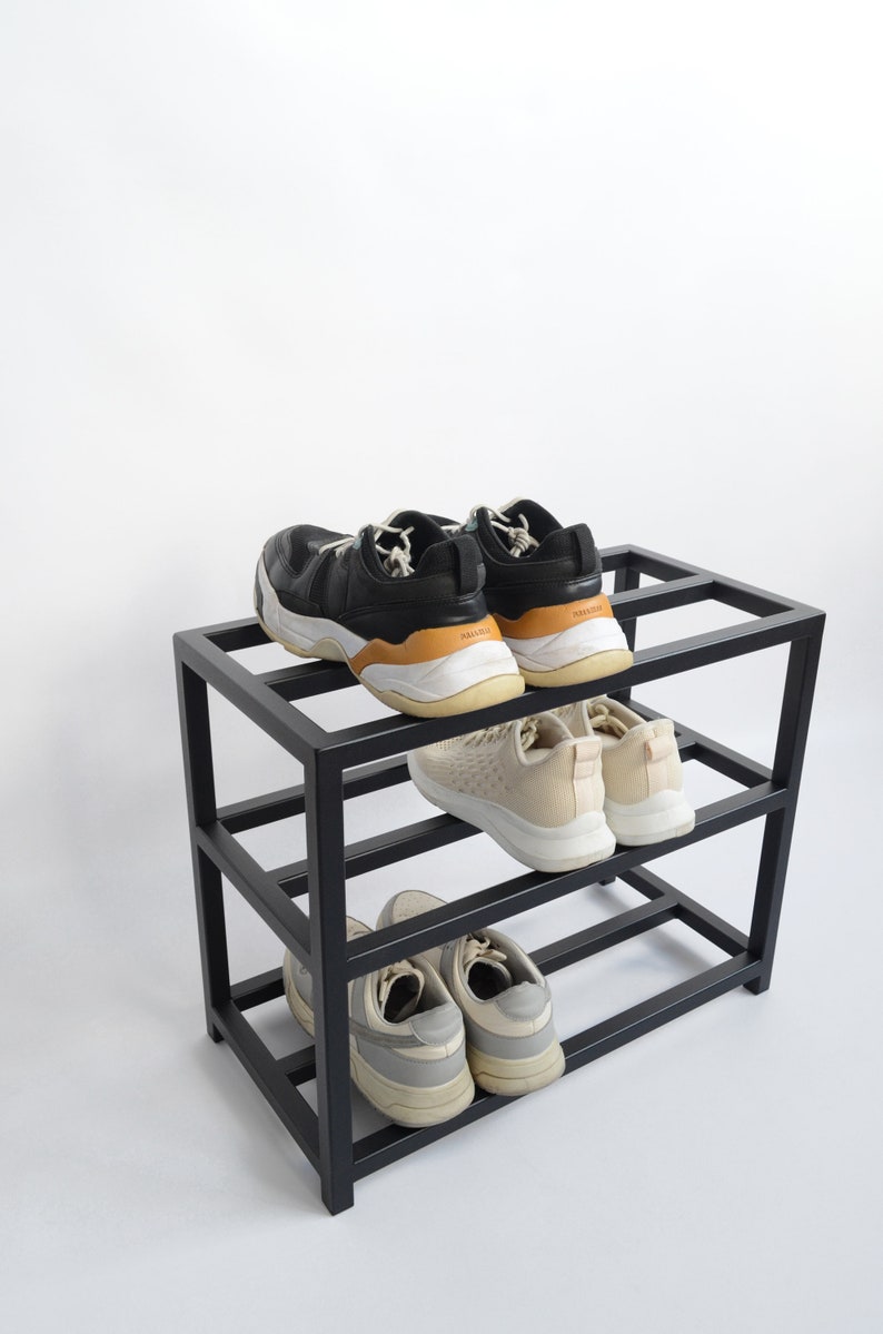 May include: Black metal shoe rack with three shelves, holding six pairs of shoes. The top shelf has two pairs of black sneakers, the middle shelf has two pairs of white sneakers, and the bottom shelf has two pairs of gray sneakers.