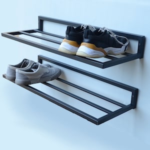 May include: Black metal shoe rack with two shelves mounted on a white wall. The top shelf has two pairs of sneakers, one black and white and one black with a brown accent. The bottom shelf has two pairs of gray sneakers.