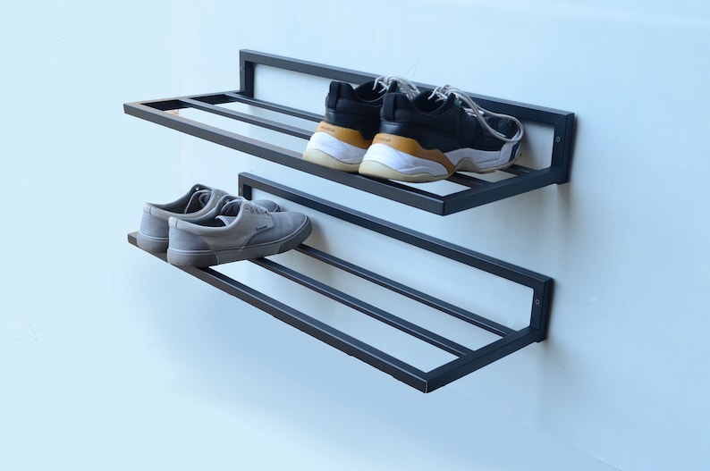 May include: Black metal shoe rack with two shelves mounted on a white wall. The top shelf has two pairs of sneakers, one black and white and one black with a brown accent. The bottom shelf has two pairs of gray sneakers.