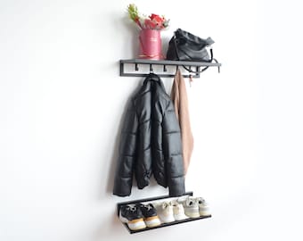 May include: Black metal wall-mounted coat rack with a shelf and hooks. A black jacket, a brown sweater, a black purse, and a bouquet of pink flowers in a red box are on the rack. A shelf below holds two pairs of white sneakers.
