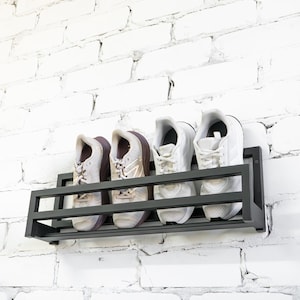 May include: A black metal shoe rack mounted on a white brick wall. The rack holds four pairs of white sneakers.