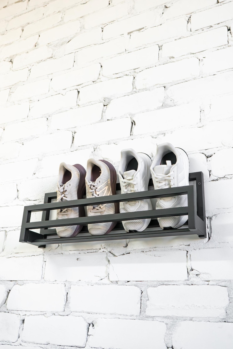 May include: A black metal shoe rack mounted on a white brick wall. The rack holds four pairs of white sneakers.