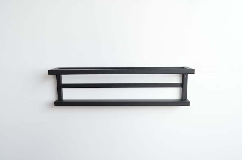 May include: A black, rectangular wall shelf with a minimalist design. The shelf has a flat base, a top frame, and two horizontal supports. The shelf is mounted on a white wall.