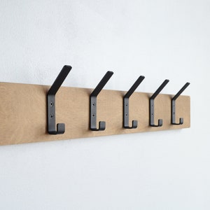 K&ouml;nnte beinhalten: Ein Wand-Kleiderhaken aus Holz mit sechs schwarzen Metallhaken. Der Haken ist aus hellbraunem Holz und hat eine nat&uuml;rliche Oberfl&auml;che.