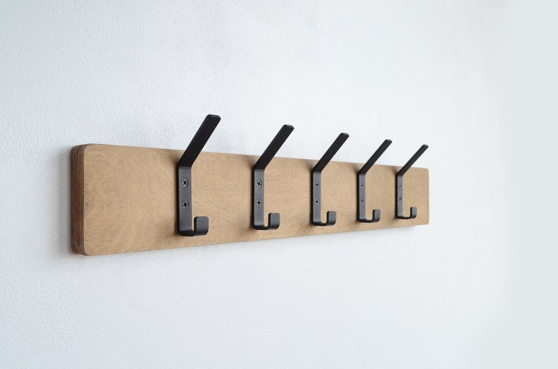 K&ouml;nnte beinhalten: Ein Wand-Kleiderhaken aus Holz mit sechs schwarzen Metallhaken. Der Haken ist aus hellbraunem Holz und hat eine nat&uuml;rliche Oberfl&auml;che.