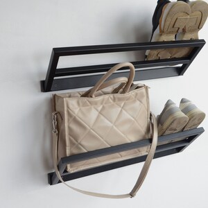 May include: Black metal wall mounted shoe rack with two shelves. A beige handbag with a long strap is hanging on the lower shelf.