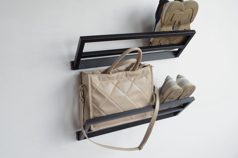 May include: Black metal wall mounted shoe rack with two shelves. A beige handbag with a long strap is hanging on the lower shelf.