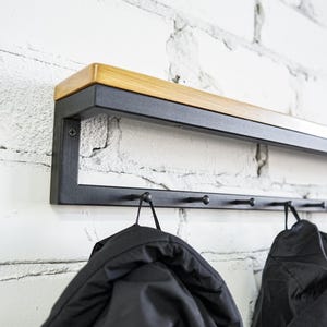 Könnte beinhalten: Eine Wandgarderobe mit einem rechteckigen schwarzen Metallrahmen und einer Ablage aus Naturholz. Die Garderobe verfügt über eine horizontale Stange mit mehreren kleinen schwarzen Haken, an denen zwei schwarze Jacken hängen.