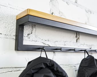 Puede incluir: Un perchero de pared con un marco rectangular de metal negro y un estante superior de madera natural. El perchero incluye una barra horizontal con varios ganchos negros pequeños, de los cuales cuelgan dos chaquetas negras.