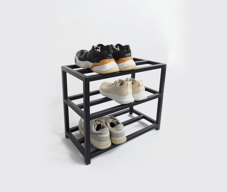 May include: A black metal shoe rack with three shelves. The top shelf has two pairs of black sneakers, the middle shelf has a pair of beige sneakers, and the bottom shelf has a pair of gray sneakers.