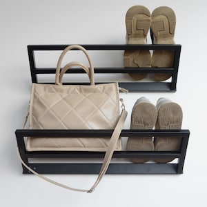 May include: Black metal wall mounted shoe and handbag rack with two shelves. A beige handbag with a long strap is on the top shelf and two pairs of brown shoes are on the bottom shelf.
